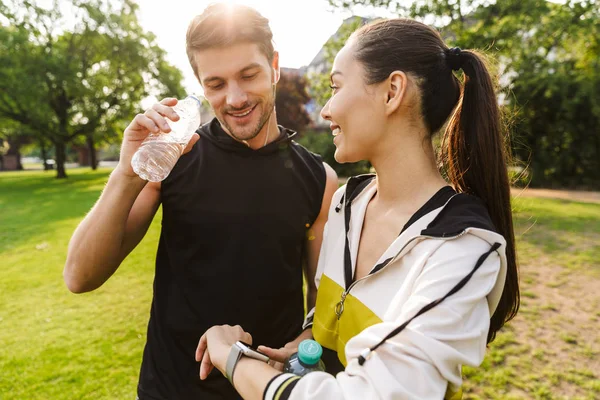 Foto de un hombre y una mujer sonrientes usando reloj inteligente y agua potable mientras hacen ejercicio en el parque de la ciudad —  Fotos de Stock