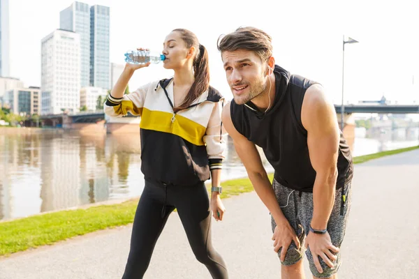 Foto de un hombre y una mujer jóvenes enfocados descansando y bebiendo agua mientras hacen ejercicio en la ribera de la ciudad —  Fotos de Stock