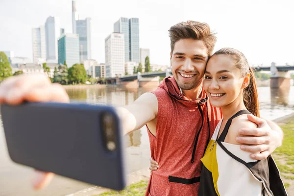 Fotó a mosolygó szép férfi és nő vesz selfie fotó mobiloddal munka közben ki a város Folyópart — Stock Fotó