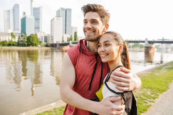 Foto de un joven feliz hombre y mujer sonriendo y abrazándose mientras se ejercita en la ribera de la ciudad — Foto de Stock