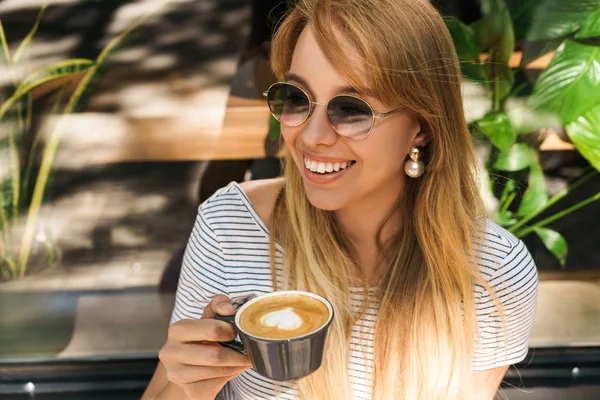 Retrato de la mujer joven de moda sonriendo y sosteniendo la taza de café mientras está sentado en la cafetería de la calle al aire libre —  Fotos de Stock