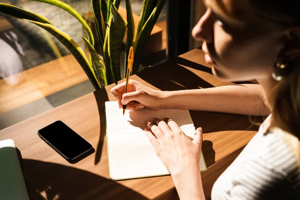 Ritratto ritagliato di bella giovane donna utilizzando prendere appunti con penna nel blocco note mentre seduto in caffè — Foto Stock