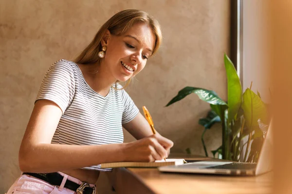 Retrato de una joven atractiva usando computadora portátil y escribiendo notas en el diario en la cafetería — Foto de Stock