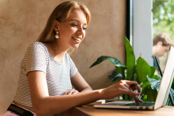 Retrato de jovem adorável usando computador portátil e escrevendo notas no livro diário no café — Fotografia de Stock