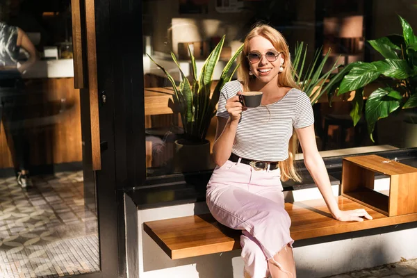 Образ задоволеної молодої жінки, яка посміхається і тримає чашку кави, сидячи у вуличному кафе на відкритому повітрі — стокове фото