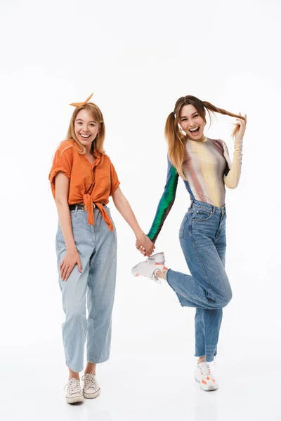 Foto de comprimento total de duas garotas adoráveis vestindo roupas coloridas sorrindo para a câmera enquanto seguram as mãos juntas — Fotografia de Stock