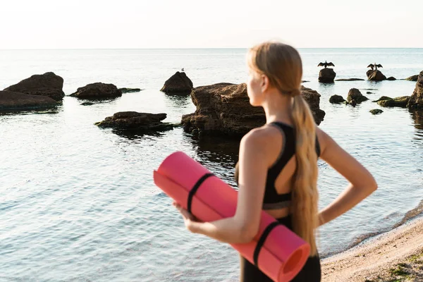 Imagem de mulher concentrada desportiva olhando para o mar e segurando tapete de ioga trabalhando fora de manhã ao ar livre — Fotografia de Stock