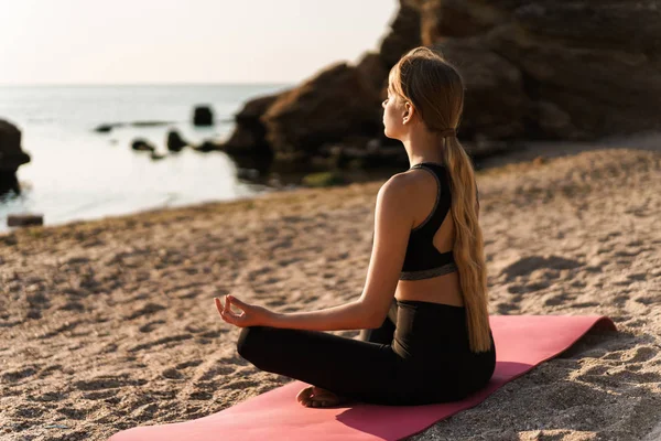 Bild av blond fokuserad kvinna mediterar medan du sitter på yogamattan vid havet på morgonen — Stockfoto