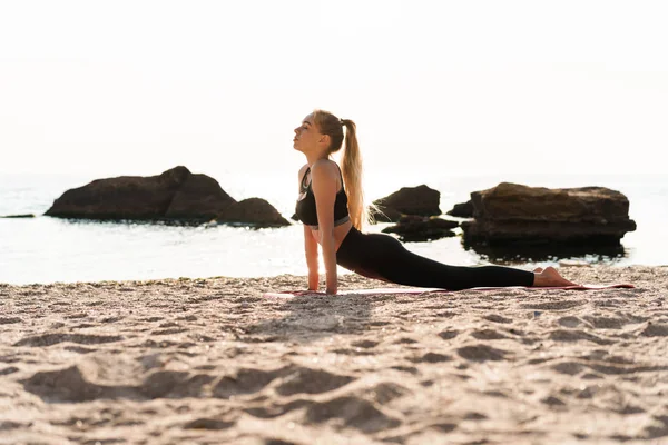 Imagen de una hermosa mujer atlética haciendo ejercicios de yoga en la estera en la playa por la mañana —  Fotos de Stock