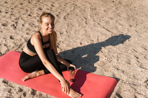 Afbeelding van lachende tevreden vrouw drinkwater na yoga oefeningen op mat aan de kust in de ochtend — Stockfoto