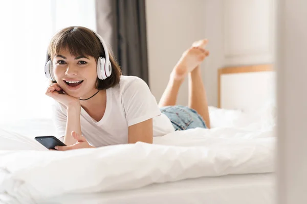 Zadowolony wesoły młody piękny kobieta wewnątrz w Dom na łóżko słuchanie muzyki rezygnować słuchawki przy pomocy zdejmowany telefon. — Zdjęcie stockowe