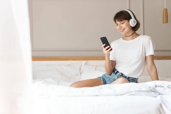 Pleased cheery young beautiful woman sitting indoors at home on bed listening music with headphones using mobile phone. — Stock Photo, Image