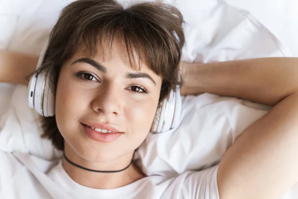 Optimistisk positiv glad ung vacker kvinna ligger inomhus hemma på sängen lyssna musik med hörlurar. — Stockfoto