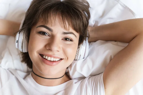 乐观积极高兴年轻美女躺在室内在家在床上用耳机听音乐. — 图库照片
