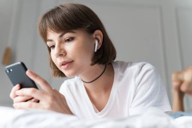 Cep telefonu kullanarak bluetooth kulaklık ile müzik dinlerken evde kapalı konsantre genç güzel kadın.