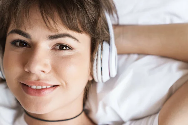 Optimiste positif heureux jeune belle femme se trouve à l'intérieur à la maison sur le lit écouter de la musique avec écouteurs . — Photo