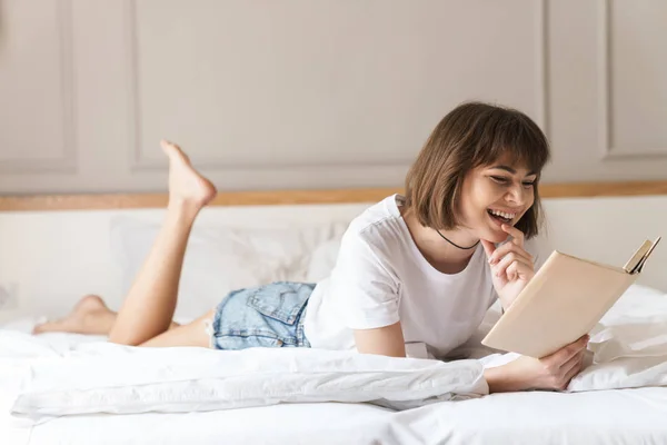 Glücklich fröhlich positive junge schöne Frau drinnen zu Hause liegt auf dem Bett Buch lesen. — Stockfoto