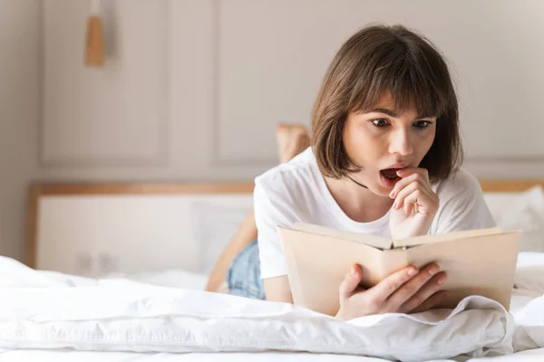 Conmocionado emocional joven hermosa mujer en el interior en casa se encuentra en la cama libro de lectura . — Foto de Stock