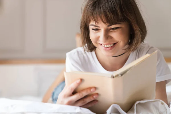 Ευτυχισμένο χαρούμενο θετικό νεαρό όμορφη γυναίκα σε εσωτερικούς χώρους στο σπίτι βρίσκεται στο κρεβάτι διαβάζοντας βιβλίο. — Φωτογραφία Αρχείου