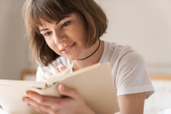 Relajante joven hermosa mujer en el interior de casa se encuentra en la cama libro de lectura . —  Fotos de Stock