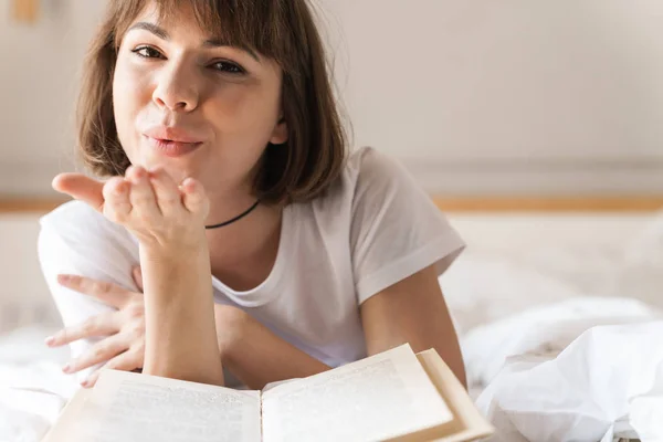 Θετική αισιόδοξη νεαρή όμορφη γυναίκα σε εσωτερικούς χώρους στο σπίτι βρίσκεται στο κρεβάτι διαβάζοντας βιβλίο ανατινάζοντας φιλιά. — Φωτογραφία Αρχείου
