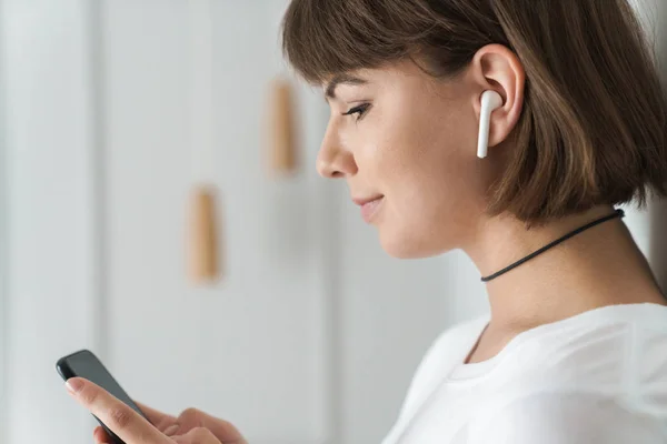 Positive young beautiful woman indoors at home listening music with bluetooth earphones using mobile phone. — Stock Photo, Image