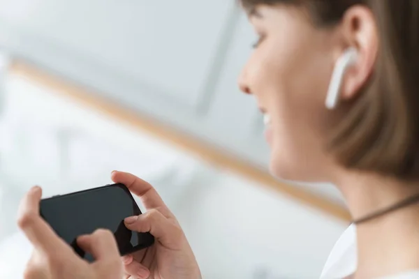 Mujer hermosa joven en el interior de casa escuchando música con auriculares bluetooth utilizando el teléfono móvil . — Foto de Stock