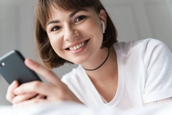 웃는 젊은 아름다운 여자는 휴대 전화를 사용하여 블루투스 이어폰으로 음악을 듣고 침대에서 집에 누워. — 스톡 사진
