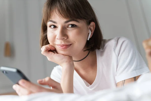 웃는 젊은 아름다운 여자는 휴대 전화를 사용하여 블루투스 이어폰으로 음악을 듣고 침대에서 집에 누워. — 스톡 사진