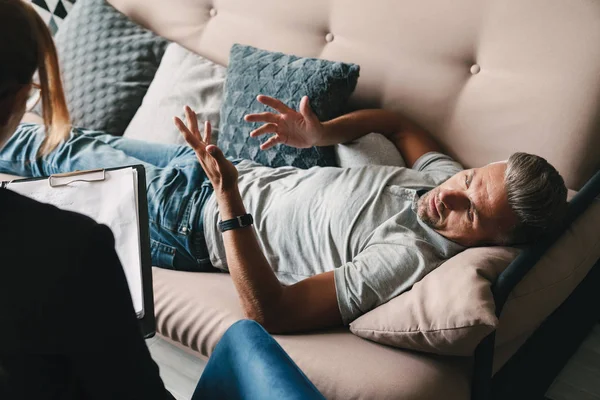 Foto de un hombre casual sin afeitar tumbado en el sofá y conversando con el psicólogo en la habitación — Foto de Stock