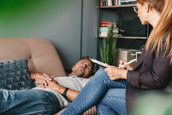 Foto de un hombre concentrado sin afeitar tumbado en el sofá y conversando con el psicólogo en la habitación — Foto de Stock