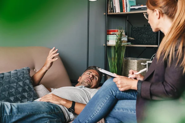 Foto de homem emocional sem barba deitado no sofá e tendo uma conversa com o psicólogo no quarto — Fotografia de Stock