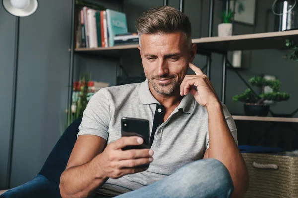 Photo of handsome caucasian man holding smartphone while sitting on armchair at home — Stock Photo, Image