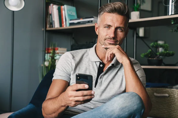 Evde koltukta otururken akıllı telefonu tutan yetişkin beyaz adamın fotoğrafı — Stok fotoğraf