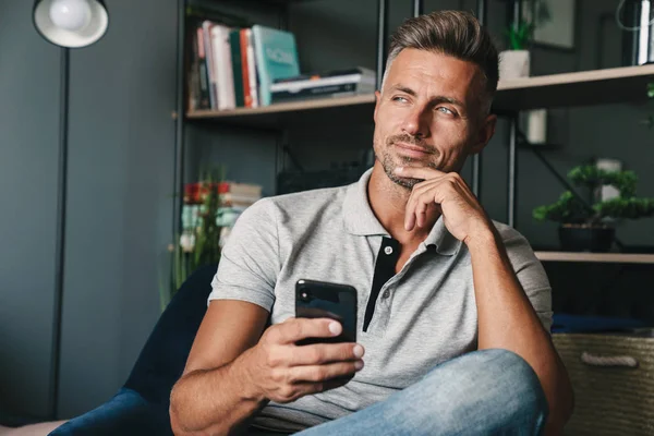 Photo of happy caucasian man holding smartphone while sitting on armchair at home — Stock Photo, Image