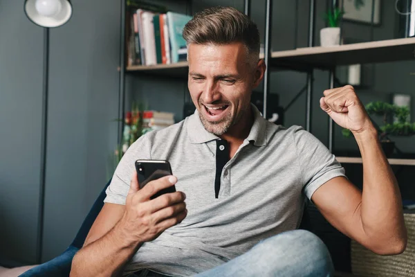 Photo of joyful caucasian man holding smartphone and rejoicing while sitting on armchair at home — Stock Photo, Image