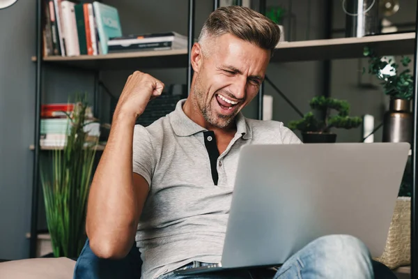 Foto de hombre adulto emocionado sonriendo y celebrando mientras usa el ordenador portátil en el apartamento — Foto de Stock