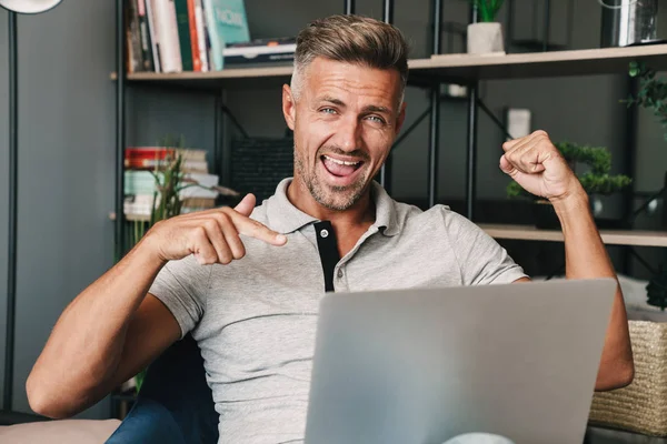Foto de hombre adulto feliz sonriendo y celebrando mientras usa el ordenador portátil en el apartamento — Foto de Stock