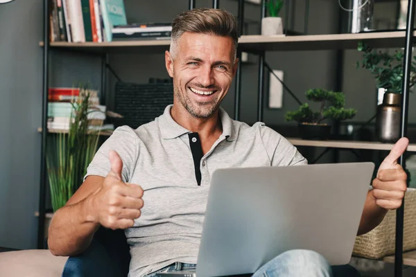 Foto de hombre adulto sin afeitar sonriendo y mostrando los pulgares hacia arriba mientras se utiliza el ordenador portátil en el apartamento — Foto de Stock