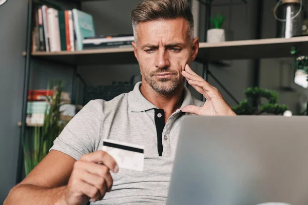 Foto de homem adulto perplexo segurando cartão de crédito e computador portátil no apartamento — Fotografia de Stock