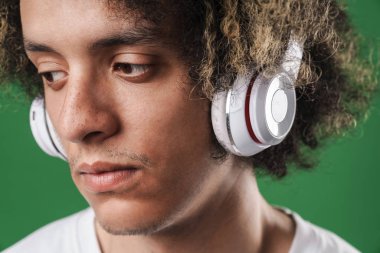 Ciddi genç kıvırcık adam kulaklık ile müzik dinleme yeşil duvar arka plan üzerinde izole poz.