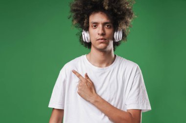 Ciddi kıvırcık adam yeşil duvar arka plan üzerinde izole poz kulaklıklar ile müzik dinleme copyspace işaret.