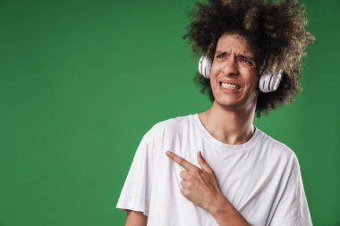 Karışık duygusal kıvırcık adam copyspace işaret kulaklık ile yeşil duvar arka plan üzerinde izole poz.