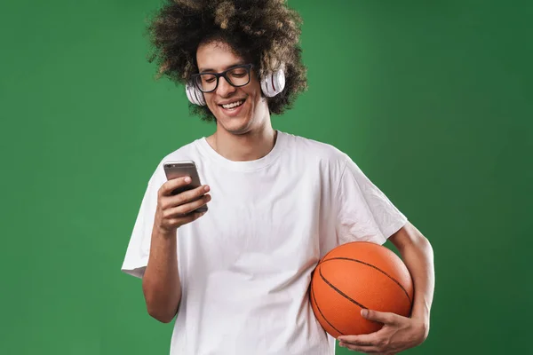 Ritratto di uomo allegro con acconciatura afro che gioca usando il cellulare e le cuffie mentre tiene il basket — Foto Stock