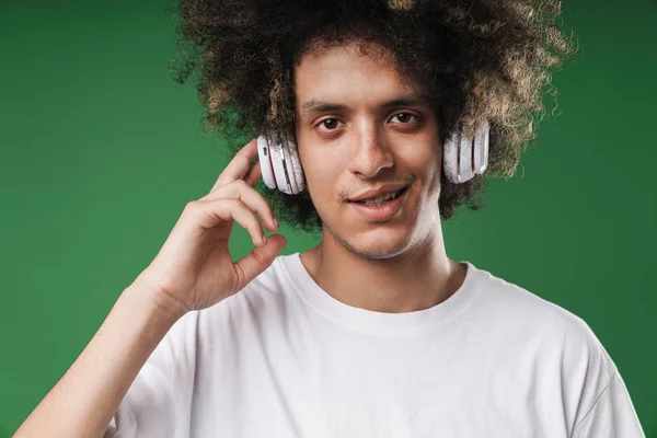 Chico rizado posando aislado sobre fondo de pared verde escuchando música con auriculares . — Foto de Stock