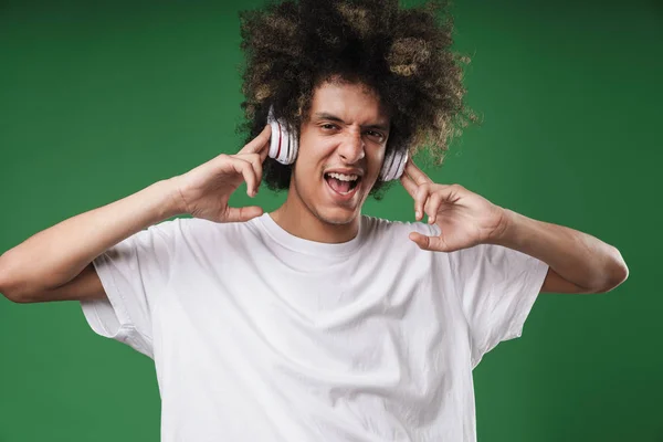 Curly Guy poserar isolerad över grön vägg bakgrund lyssnar musik med hörlurar. — Stockfoto