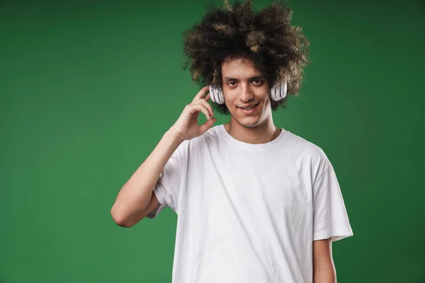 Krullend vent poseren geïsoleerd over groene muur achtergrond luisteren muziek met koptelefoon. — Stockfoto