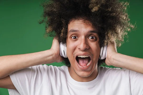 Geschokt schreeuwen jonge krullend Guy poseren geïsoleerd over groene muur achtergrond luisteren muziek met koptelefoon. — Stockfoto