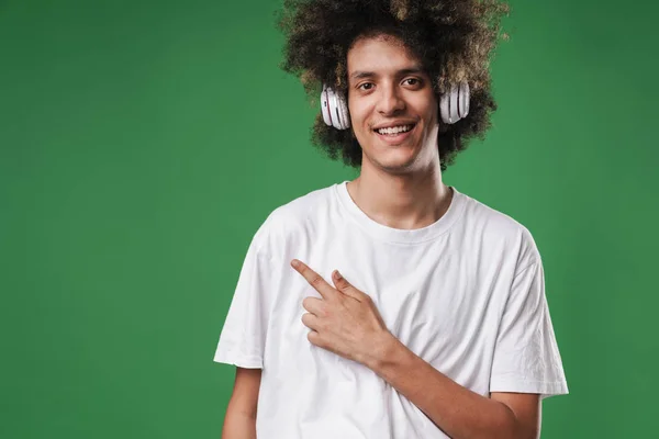 Glimlachende krullend man poseren geïsoleerd over groene muur achtergrond luisteren muziek met koptelefoon wijzend naar copyspace. — Stockfoto