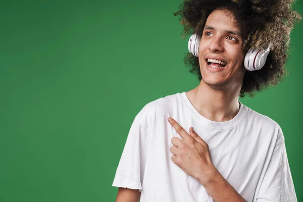 Sorrindo cara encaracolado posando isolado sobre fundo parede verde ouvir música com fones de ouvido apontando para copyspace . — Fotografia de Stock